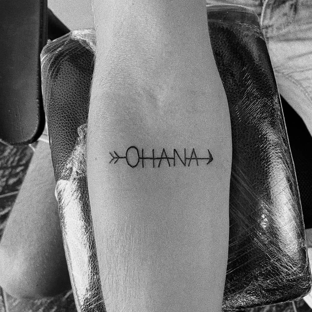 Simple black tattoo of the word 'Ohana' with arrows on each side, symbolizing family, direction, and unity.
