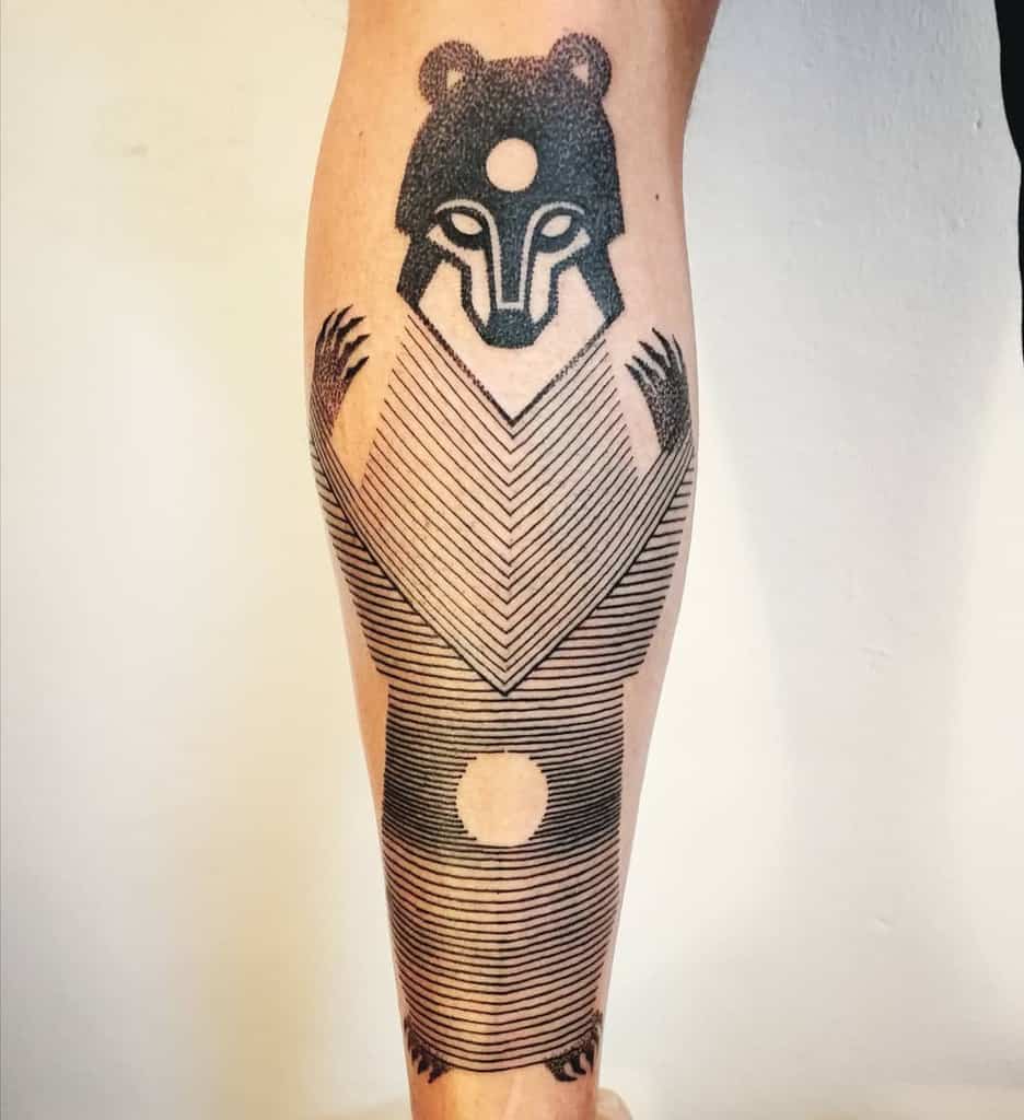 Tattoo of a stylized bear with geometric lines and circular symbols, inked in black on the lower leg.