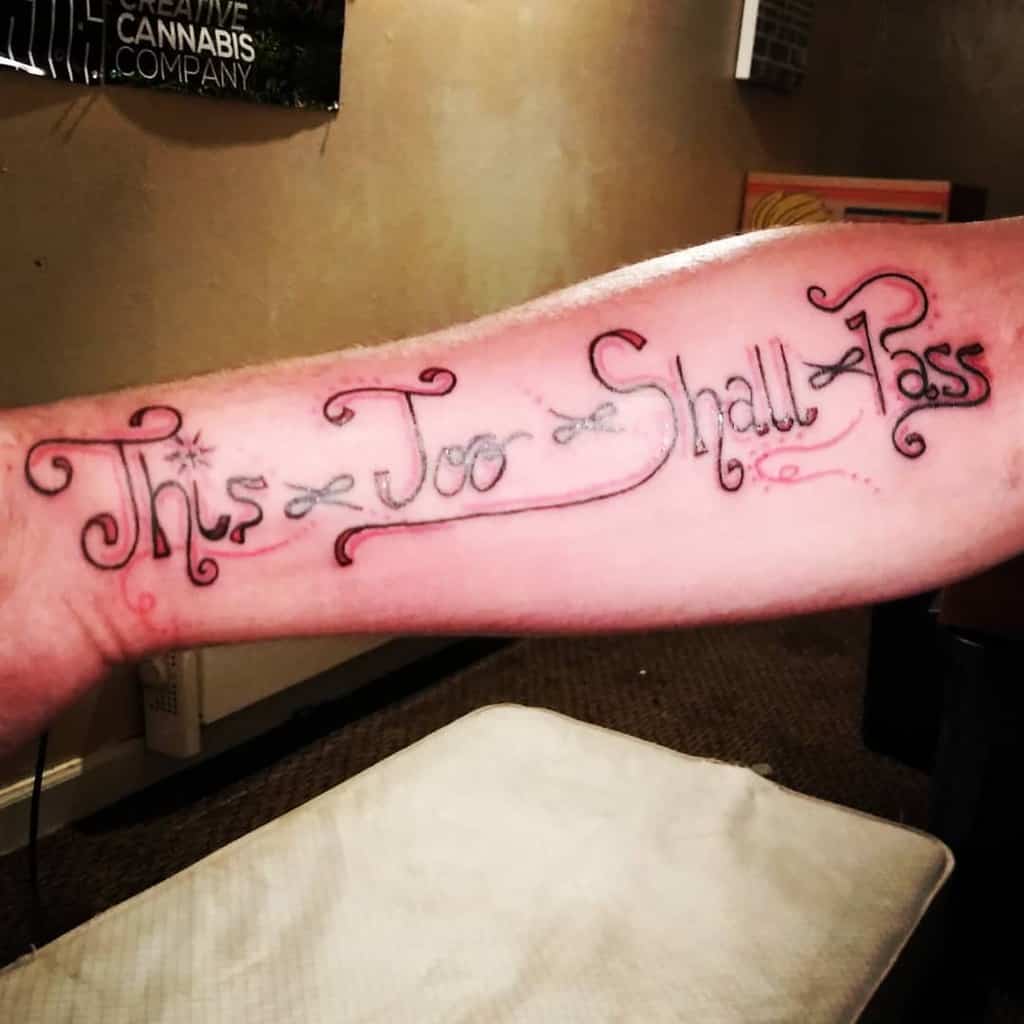 A "This Too Shall Pass" tattoo on a person's forearm, with intricate swirls and decorative elements surrounding the text.