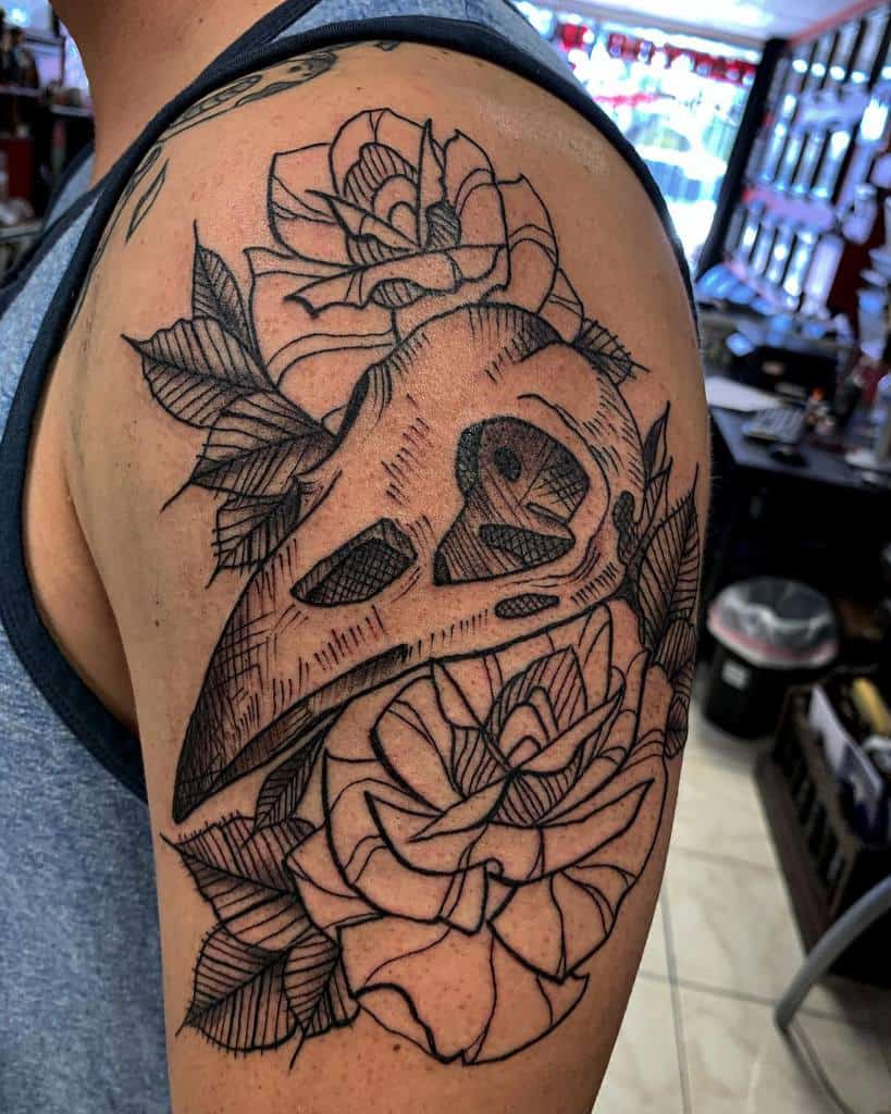 Black ink tattoo of a detailed raven skull surrounded by roses, featuring bold linework and shading on the upper arm.
