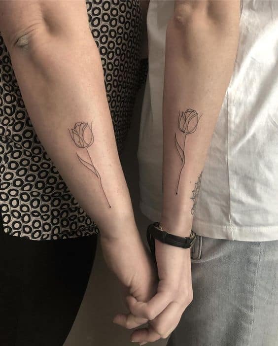 Matching minimalist tulip tattoos on forearms, featuring delicate line art flowers with curved stems, as two people hold hands.
