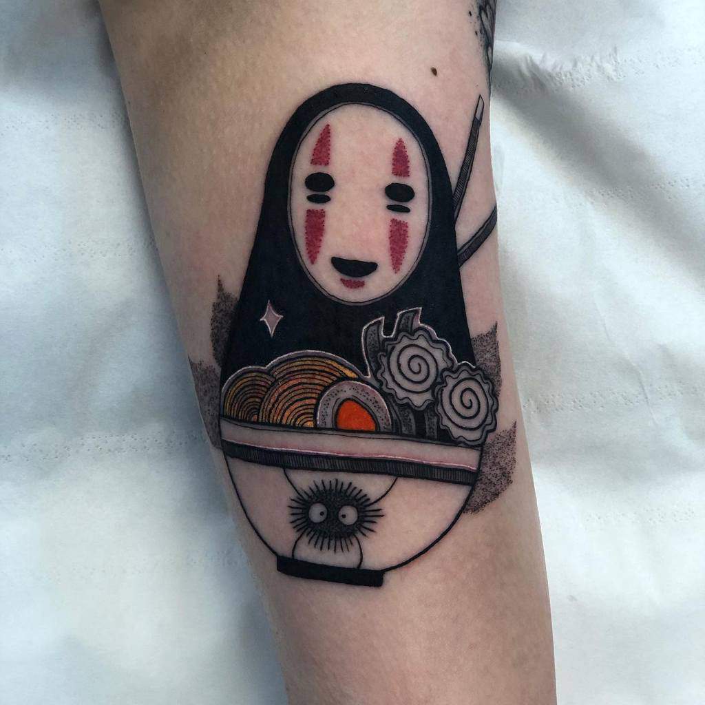Playful Spirited Away tattoo of No-Face inside a ramen bowl, with chopsticks, narutomaki, and a soot sprite peeking from the bowl’s design