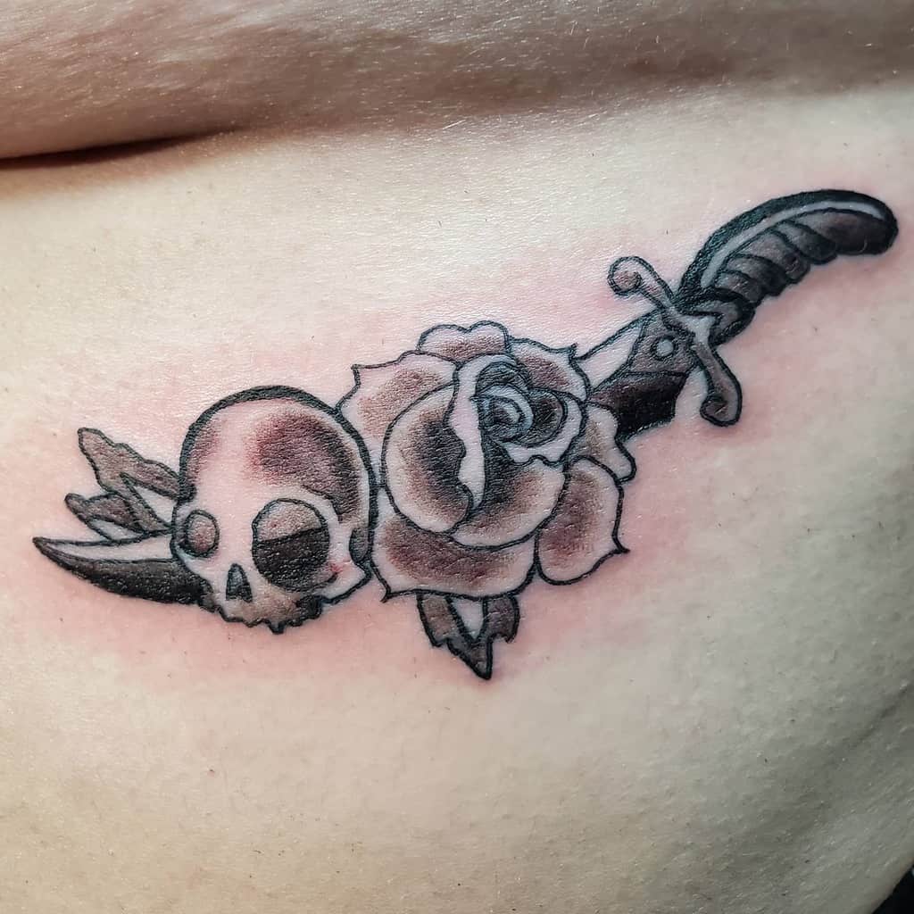 Black and grey tattoo of a dagger piercing through a skull and rose, symbolizing strength and mortality, inked on the side torso.