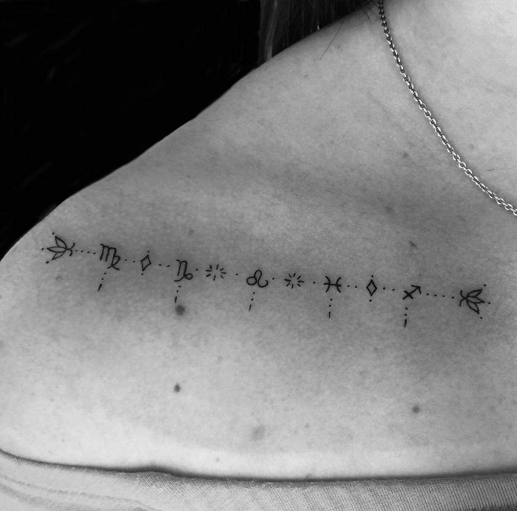 Minimalist zodiac symbol tattoo across the collarbone, featuring fine-line astrology signs connected by delicate dots.