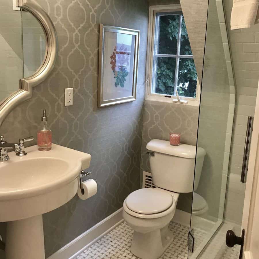 Small bathroom with gray patterned wallpaper, pedestal sink, and framed window.
