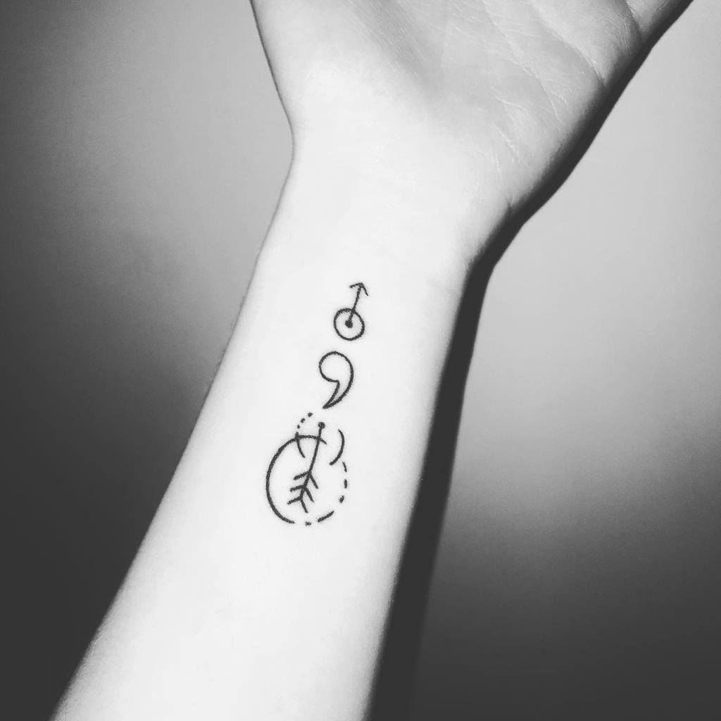 The tattoo features a unique arrangement of symbols, including a semicolon, a male symbol, and an abstract circle with leaf-like elements, creating a meaningful design on the inner forearm.