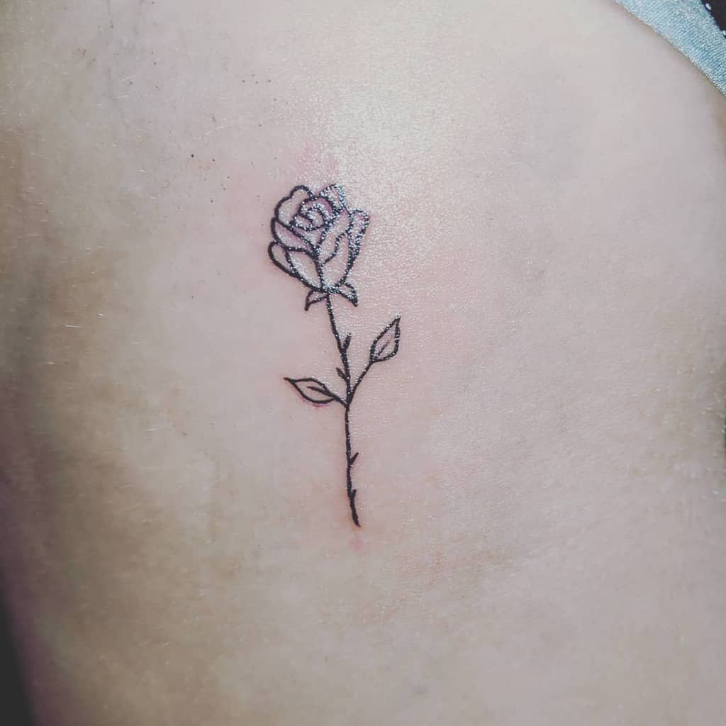 A simple black outline of a rose tattoo elegantly graces the skin, with a delicate stem that adds an extra touch of elegance