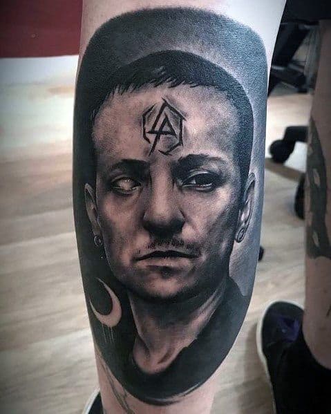 Realistic black and gray tattoo of Chester Bennington, featuring the Linkin Park logo on his forehead and a crescent moon on the leg.