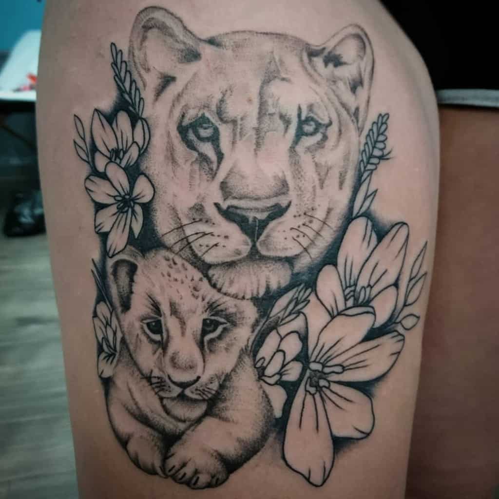 Black and grey thigh tattoo featuring a lioness and cub surrounded by floral details, symbolizing strength, protection, and motherhood.