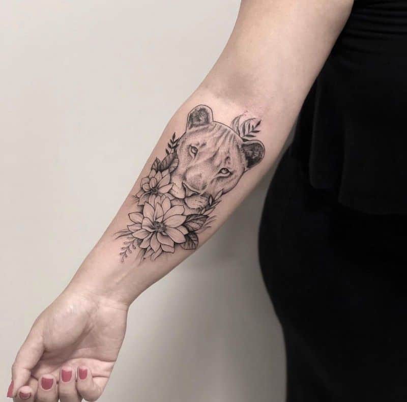 Delicate black and grey lioness tattoo on the forearm, featuring floral details and fine shading for a soft yet fierce look.