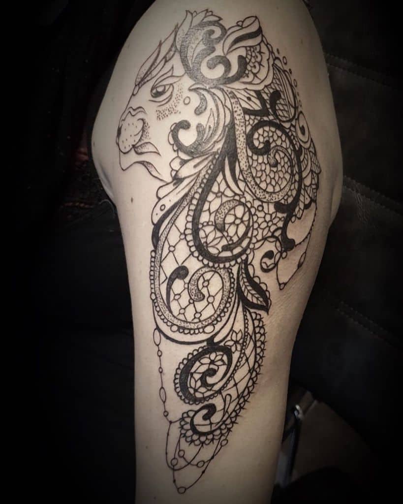 Black ink upper arm tattoo of a lion with an intricately detailed mane featuring lace patterns, ornamental filigree, and beaded chain accents.