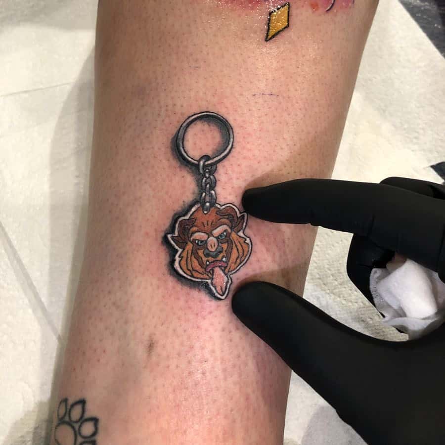 Realistic tattoo of a Beast keychain from Beauty and the Beast, designed with shading to create a 3D effect on the ankle.
