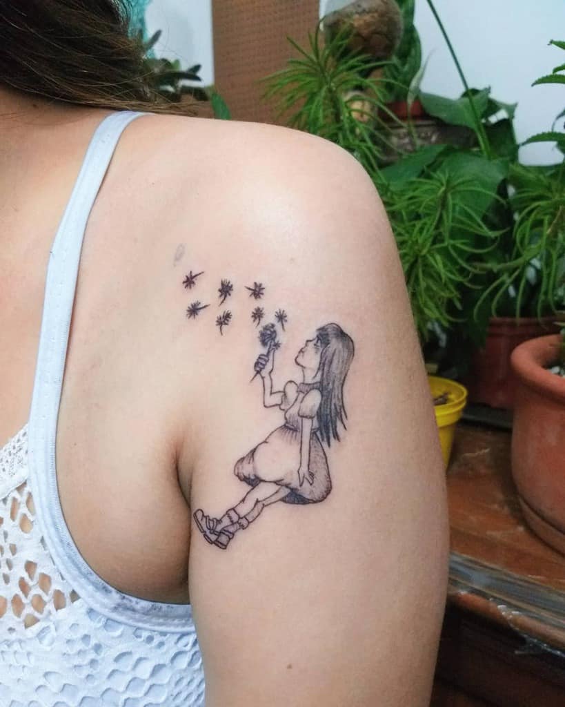 A tattoo of a girl sitting and blowing dandelion seeds, with the seeds drifting away, on the upper arm, surrounded by plants.