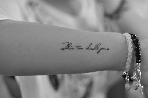"This too shall pass" tattoo in cursive on the arm with bracelets visible.