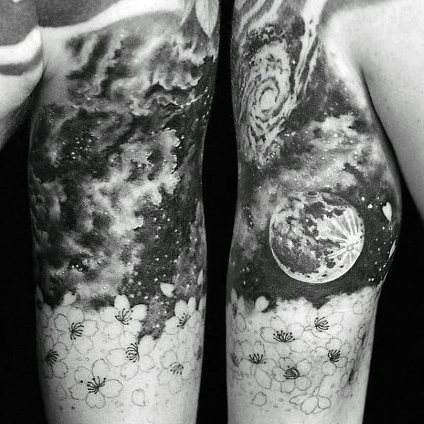 Black and white tattoos of galaxies, moon, and flowers on inner arms