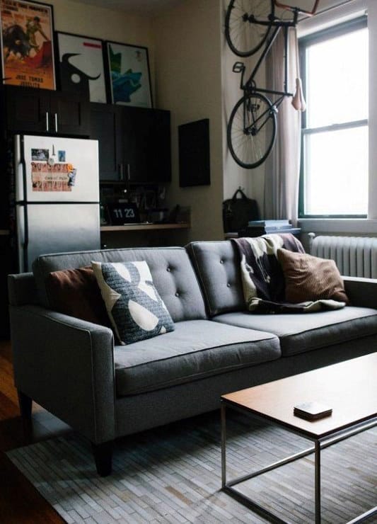 Small living room with gray sofa, wall-mounted bike, and compact kitchen in background.