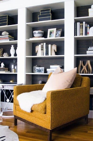 A cozy room with a mustard yellow armchair, a white pillow, and bookshelves filled with decor items and books