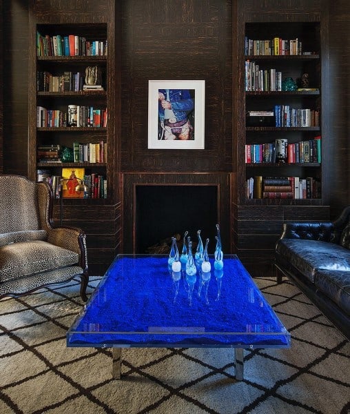 Cozy living room with a blue glass table, leather and patterned chairs, bookshelves, and abstract art above fireplace