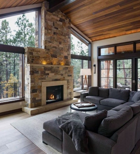 Cozy living room with vaulted wood ceiling, stone fireplace, large windows, and a modern gray sectional sofa.