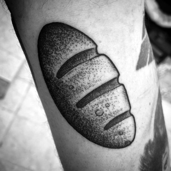 Black ink tattoo of a rustic bread loaf with bold dotwork shading and deep slashes, creating a textured and artistic design on the skin.