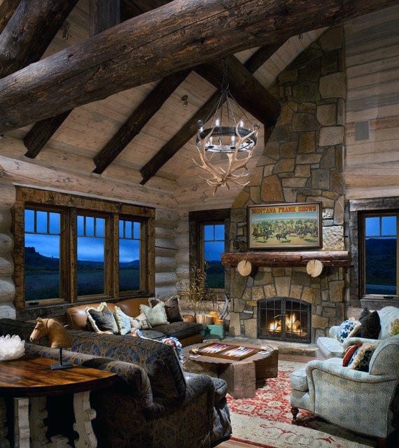 Rustic log cabin living room with stone fireplace, vaulted ceiling, and antler chandelier.