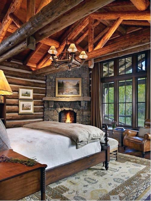Rustic log cabin bedroom with stone corner fireplace, wood beams, chandelier, and leather armchair.