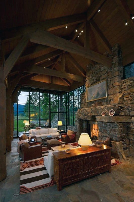 Modern log cabin living room with stone fireplace, large windows, and vaulted wooden ceiling.