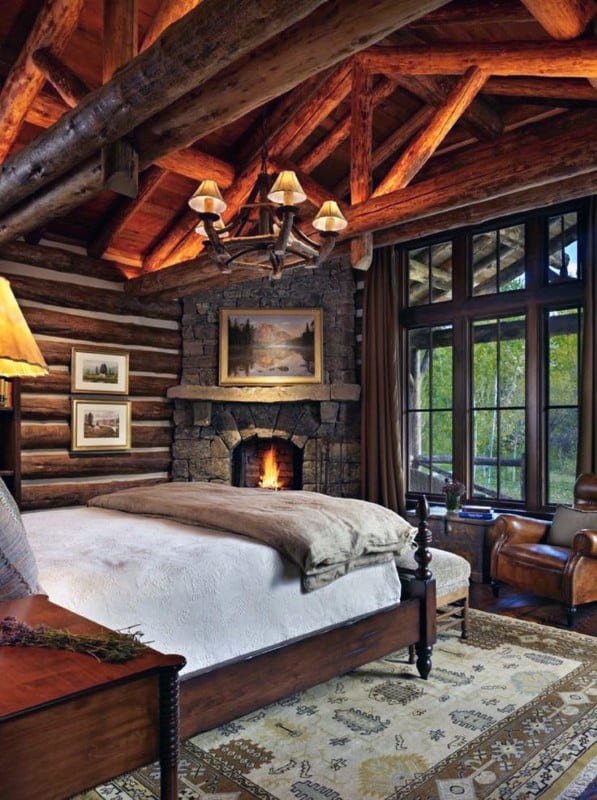 Cozy log cabin bedroom with stone fireplace, wooden beams, and large windows with scenic views.