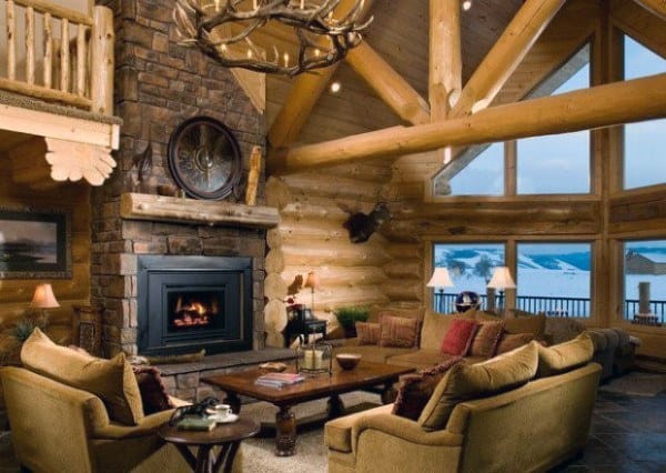 Cozy log cabin living room with large windows, stone fireplace, antler chandelier, and comfortable seating.