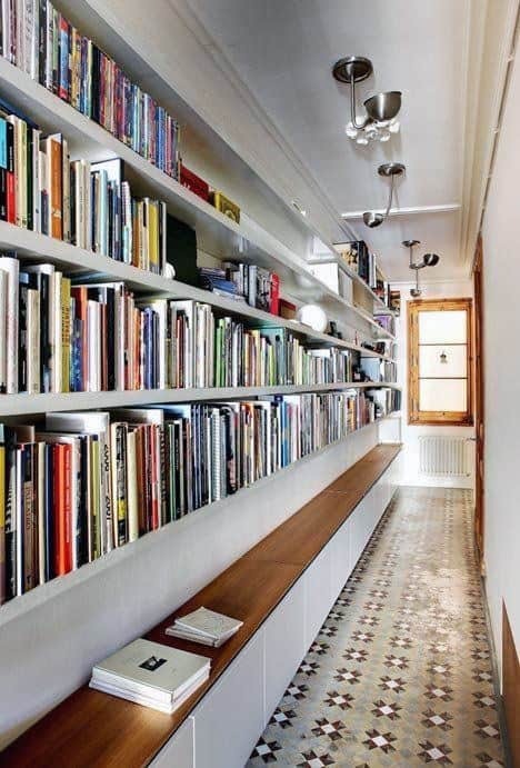 A hallway resembling a library with wall-mounted bookshelves, a long wooden bench, and a patterned tile floor