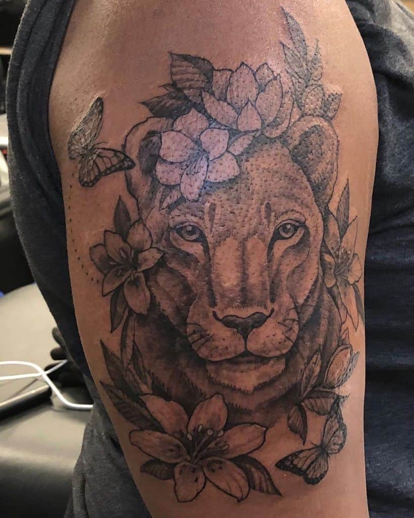 Black and grey lioness tattoo on the upper arm, featuring floral elements, butterflies, and intricate shading for a bold yet elegant look.
