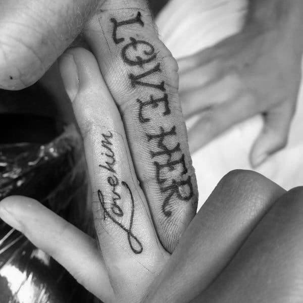 Matching inner finger tattoos that say "Love Him" and "Love Her," symbolizing devotion and a deeply personal commitment between partners.