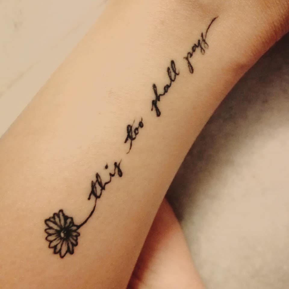 A "This too shall pass" tattoo in flowing cursive, seamlessly extending from a small daisy, symbolizing resilience and the passage of time.