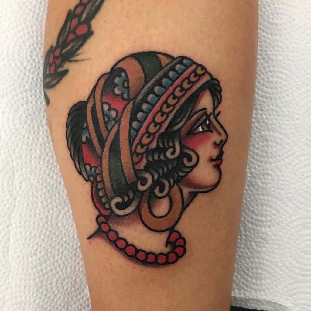 Traditional tattoo of a gypsy woman in profile with a colorful headscarf, gold earrings, and a red beaded necklace on the leg.