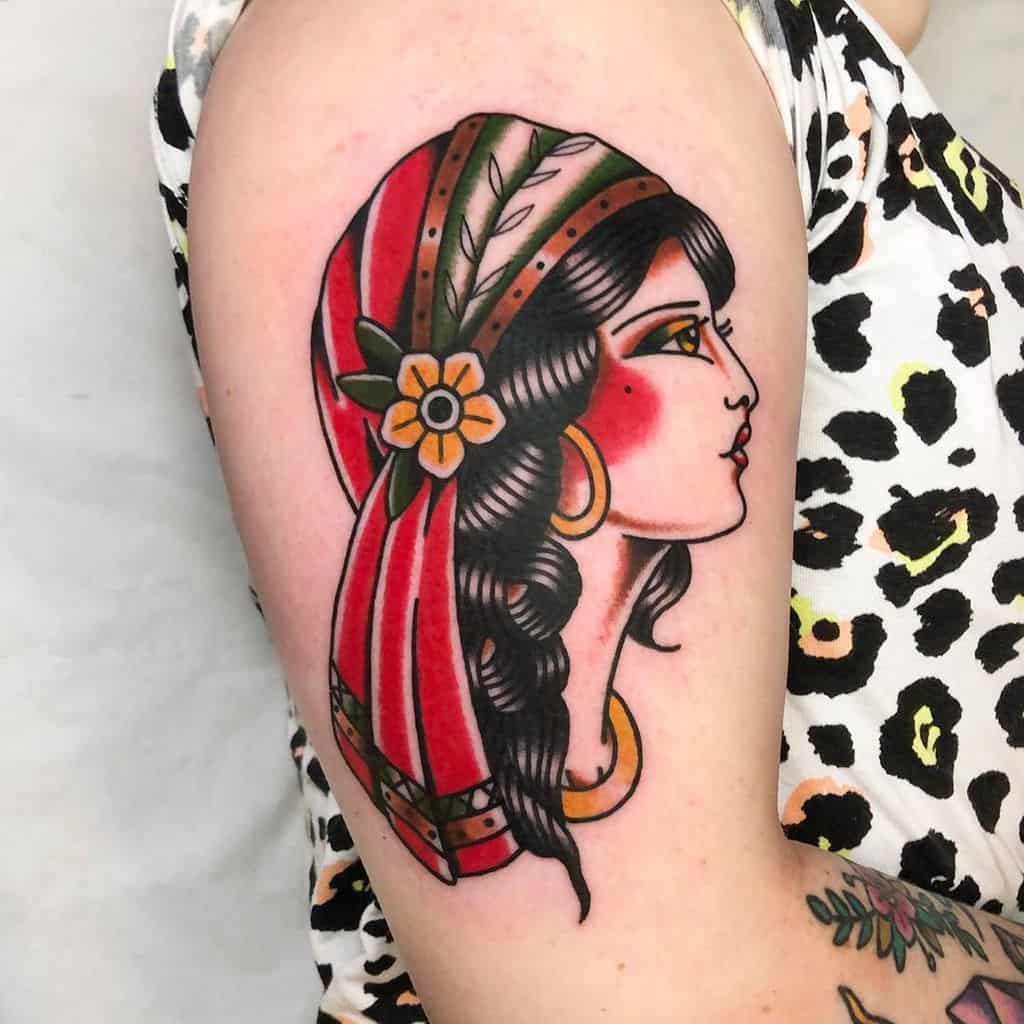 Bold traditional tattoo of a gypsy woman with a red headscarf, braided hair, and gold jewelry on the upper arm.