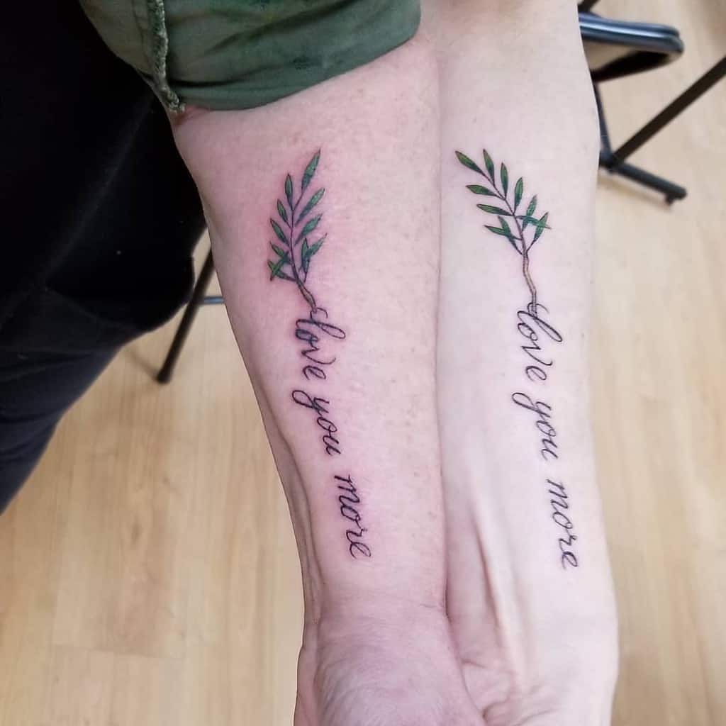Matching sister tattoos featuring a delicate olive branch with the phrase 'love you more' in cursive, symbolizing a deep bond