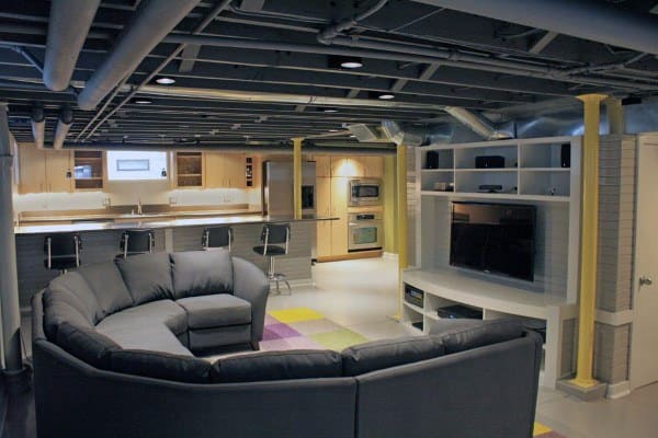 Industrial-style basement with exposed ceiling pipes, sectional sofa, and modern kitchen area.