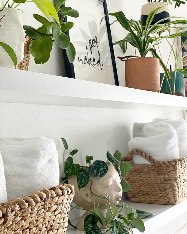 White bathroom shelves with potted plants, wicker baskets, and rolled towels.
