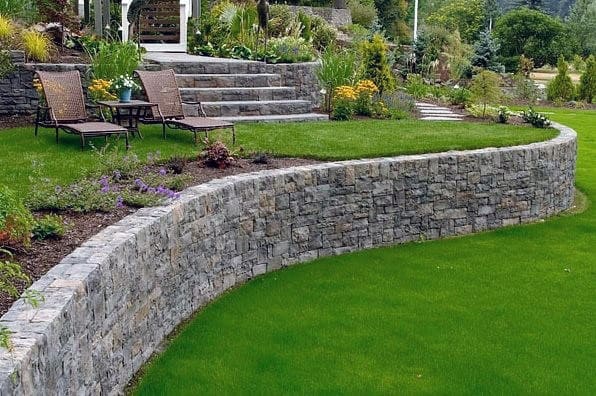 Curved stone retaining wall with a lush green lawn, garden beds, and two reclining chairs with a side table.