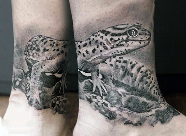 Realistic black and grey gecko tattoo wrapped around the ankle, featuring detailed textures, spots, and rocky background shading.