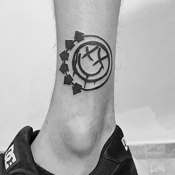 Black and white tattoo of the Blink-182 smiley face logo with arrows, placed on the lower leg near the ankle.