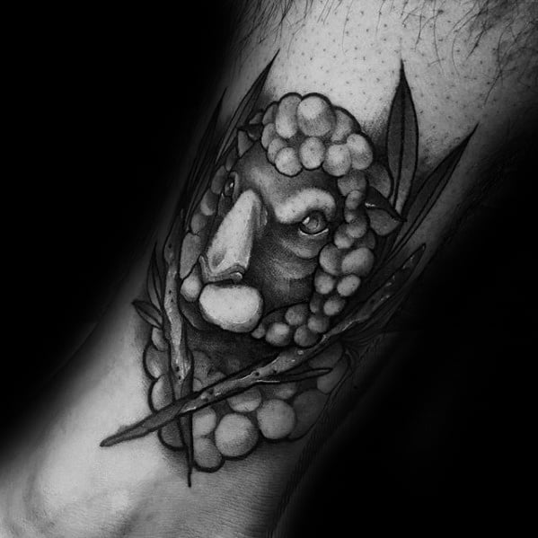 Realistic black and gray sheep head tattoo on the leg, surrounded by leaves and detailed shading.