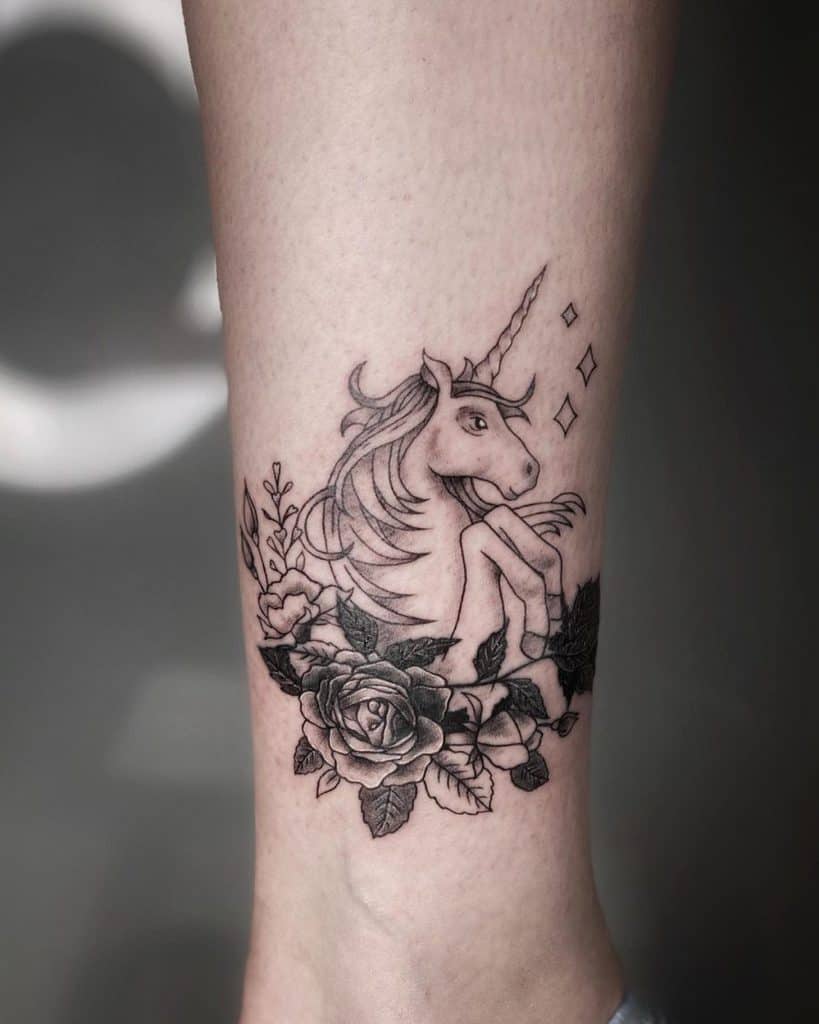 Elegant black ink tattoo of a unicorn sitting among detailed roses and leaves, with sparkling accents for a whimsical and mystical touch.