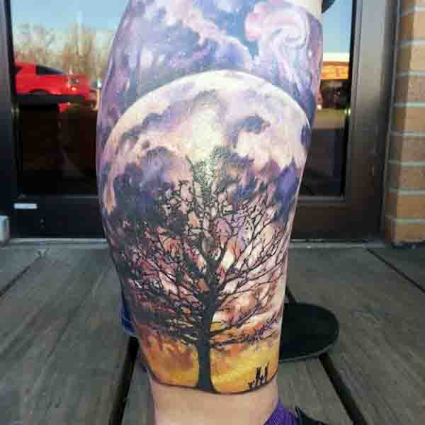 Tattoo on leg of a full moon behind a tree silhouette with vibrant purple and orange hues