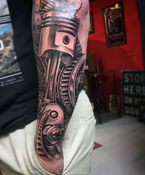 Forearm tattoo of mechanical gears and a piston, depicted in black and grey ink