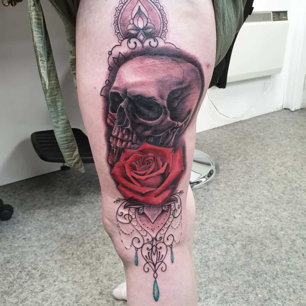 Elegant leg tattoo featuring a dark skull, a vibrant red rose, and intricate ornamental details with jewel-like accents.