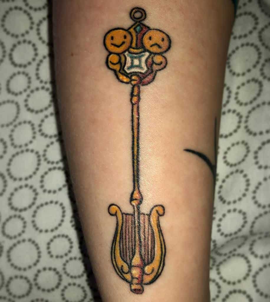 Colorful tattoo of Lucy Heartfilia's Gemini Celestial Spirit Key from Fairy Tail, featuring intricate gold details and expressive twin faces.