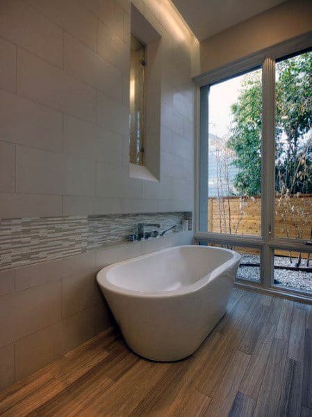 Freestanding tub with textured tile accent, wood-look floor, and large window view.