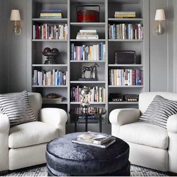 Cozy reading nook with two armchairs, a round ottoman, and a bookshelf filled with books in a stylish, gray room