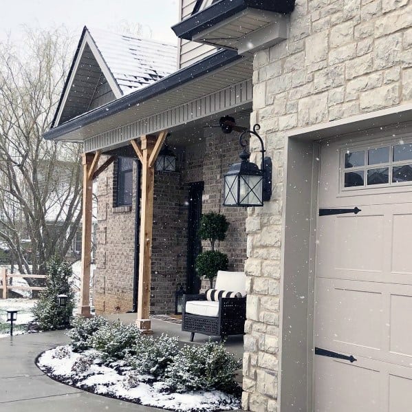 Cozy home with stone cladding, wooden porch posts, black lantern lighting, and a snow-dusted landscape for a warm winter look.